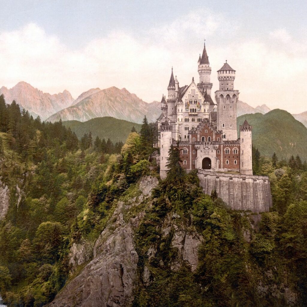 Auf dem Bild ist das Schloss Neuschwanstein zu sehen. Es steht auf einem hohen Felsen und ist von einem grünen Mischwald umgeben. Im Hintergrund kann man die hohen Berge der Voralpen sehen. 