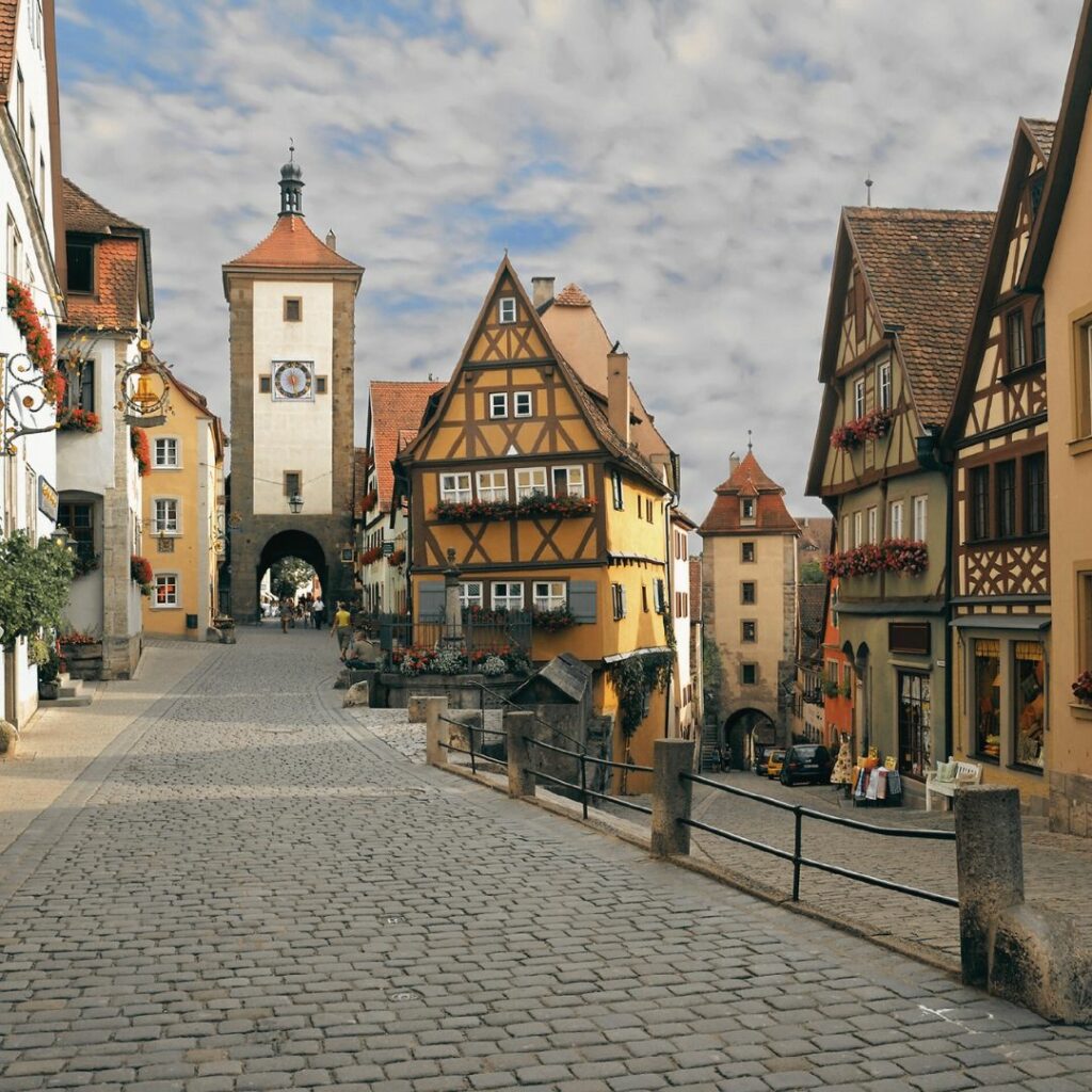 Das Bild zeigt eine berühmte Straße in der historischen Altstadt in Rothenburg ob der Tauber. Zu sehen sind alte Fachwerkhäuser und ein Turm mit einem Turmbogen. Der Boden ist mit Pflastersteinen gepflastert. Die Straße ist ganz leer und viele Blumen hängen an den Fenstern. 