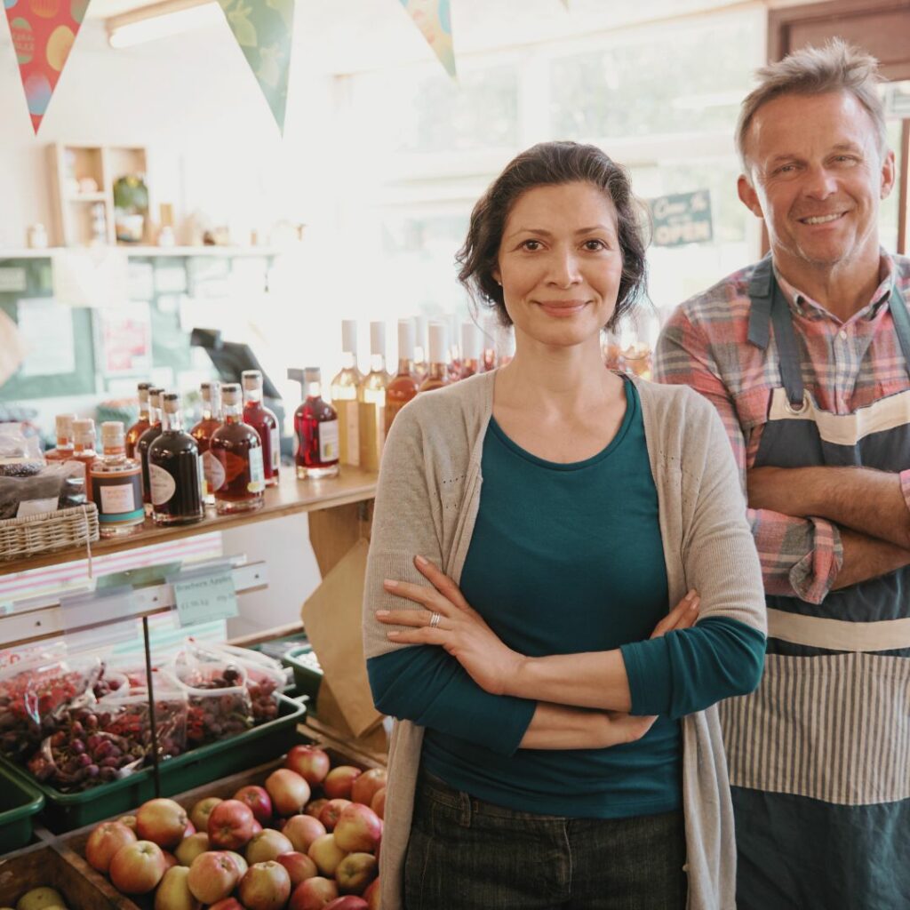 Eine Bäuerin und Ein Bauer stehen in ihrem Hofladen. Im Hintergrund sieht man verschiedene Produkte wie Äpfel, Kirschen und selbstgemacht Liköre. 
