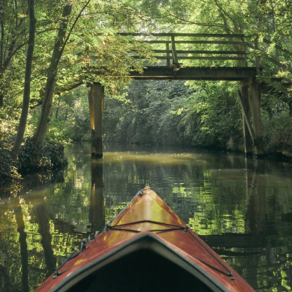 In dem Bild fährt eine Person mit einem Kajak über einen ruhigen Fluss oder Kanal im Spreewald. das Wasser ist leicht grün gefärbt und die Bäume am Ufer spiegeln sich im Wasser. Es ist dicht mit Bäumen und Sträuchern bewachsen. Eine kleine Holzbrücke führt über den Kanal und verbindet so beide Ufer.