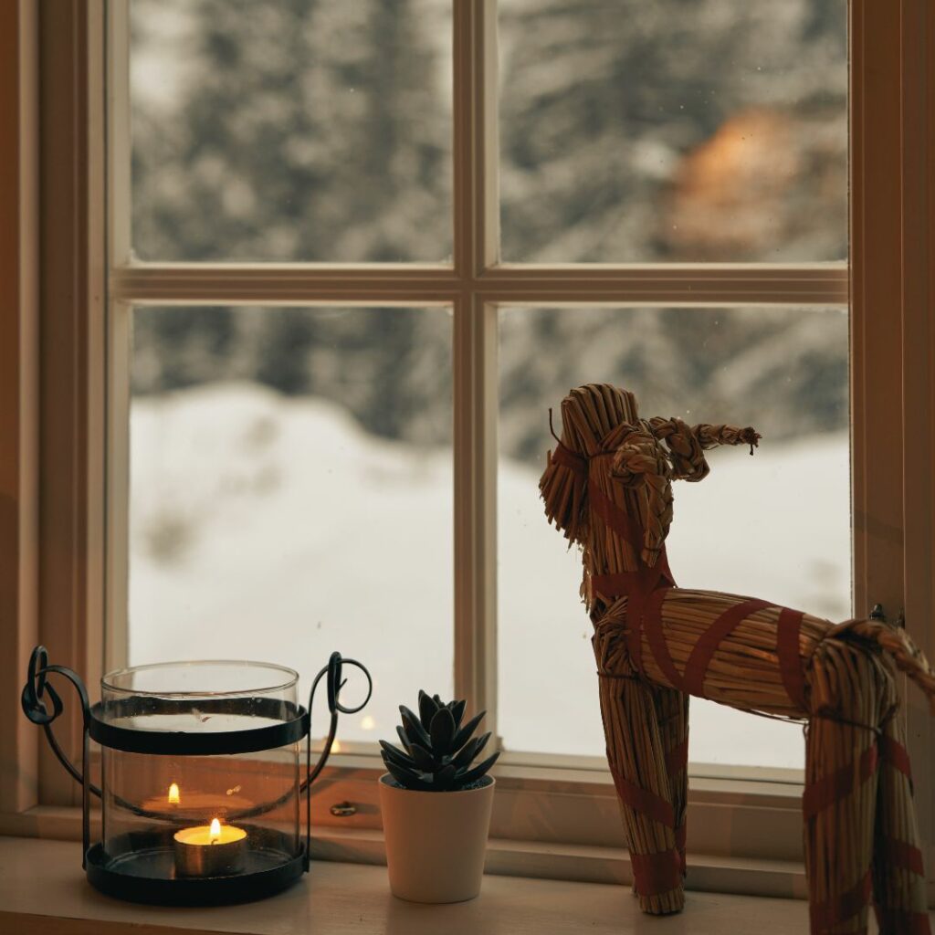 In einem alten Holzfenster stehen eine Kerze, eine kleine Sukkulente und ein skandinavischer Julbock. Draußen ist es kalt und es liegt Schnee. Die Kerze brennt und strahlt ein warmes Licht aus.
