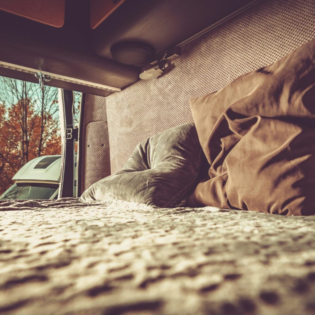 Im Heck eines Camper Vans befindet sich ein Bett, auf dem kuschelige Decken und Kissen liegen. 
