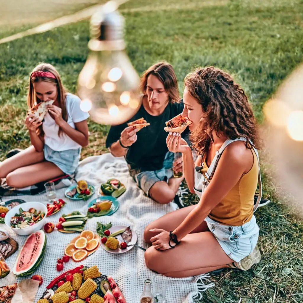Eine Gruppe von Freunden sitzt auf einer Decke. Auf der Decke befinden sich viele verschiedene Lebensmittel wie Obst und Gemüse. Sie essen gemeinsam und sitzen neben der Decke. Sie werden von der warmen Sonne angescheint. 