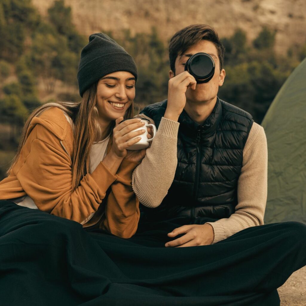 Eine Frau und ein Mann trinken Getränke aus Camping-Tassen. Die Frau lächelt und hält ihre Hände fest an der Tasse fest. Der Mann trinkt aus der Tasse. 