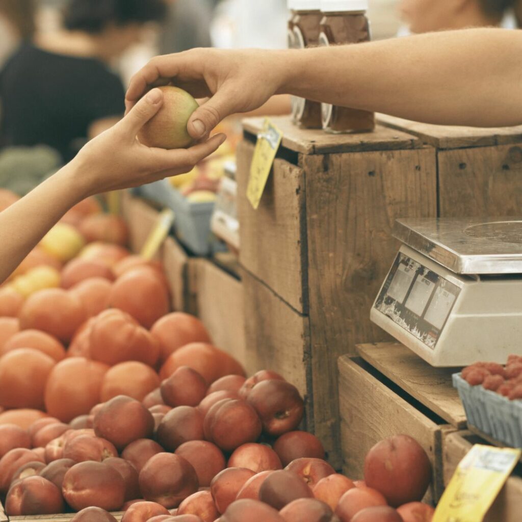 An einem Wochenmarktstand übergibt ein Verkäufer einen Apfel an einen Käufer. 